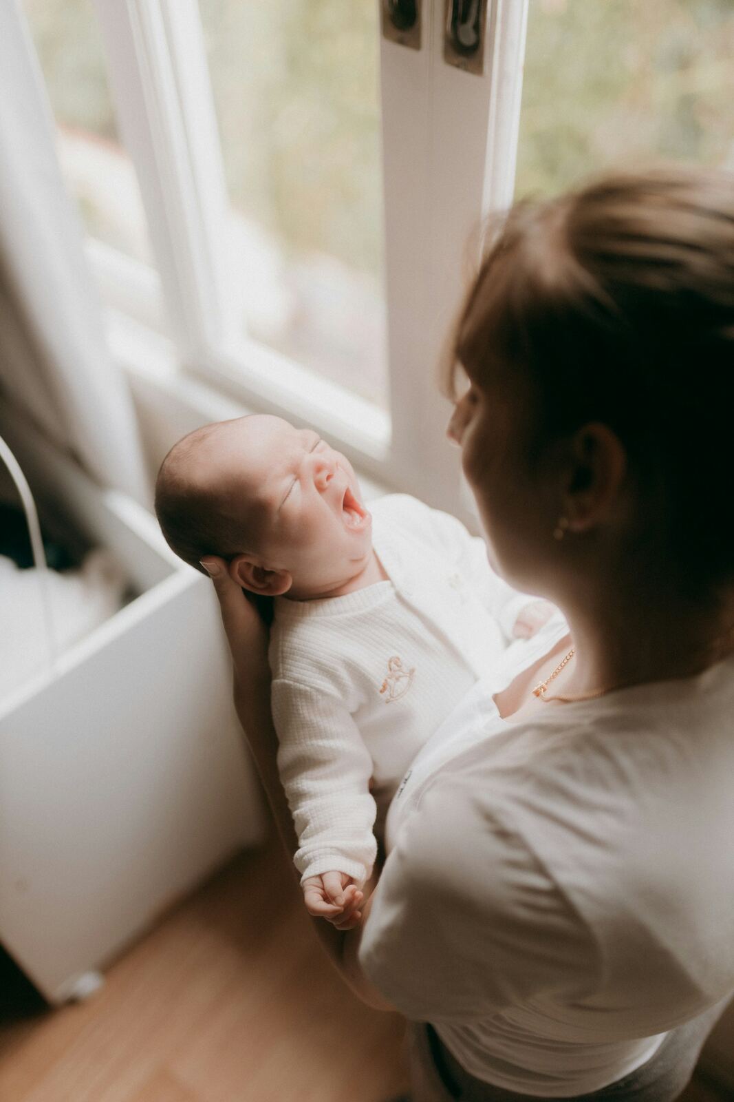 Mãe segura o bebê no colo enquanto ele boceja, em luz de manhã suave e quente